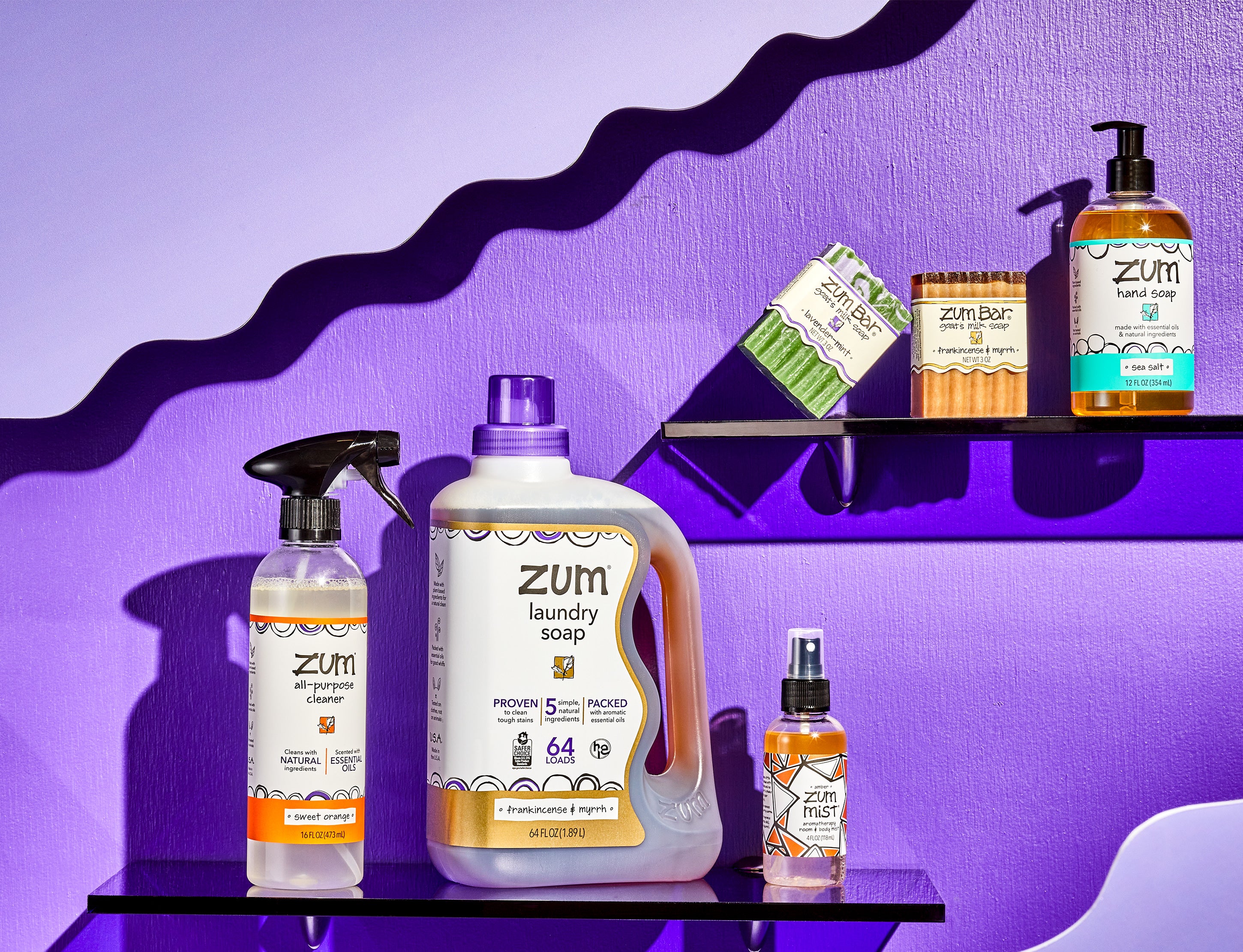 Sweet-Orange All-Purpose Cleaner bottle with a sprayer, Frankincense & Myrrh Laundry Soap bottle, and Amber mist bottle with a sprayer on a purple shelf on the bottom left half of the image. Lavender-Min and Frankincense & Myrrh labeled bar soaps and Sea Salt Hand Soap bottle with a pump on a purple shelf on the top right half of the image. Purple wood background with light purple wavy designs coming from the corners on the top left and bottom right.