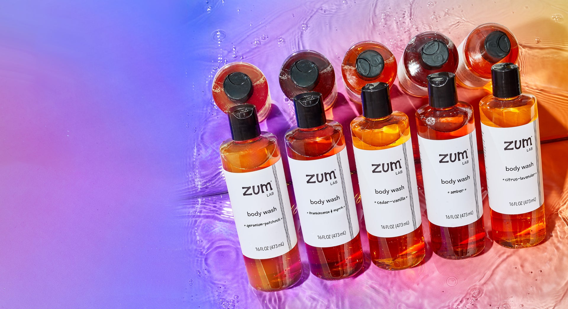 Geranium-Patchouli, Frankincense & Myrrh, Cedar-Vanilla, Amber, and Citrus-Lavender scented body wash bottles lined up against a mirror on a watery surface. Purple to orange gradient background color.
