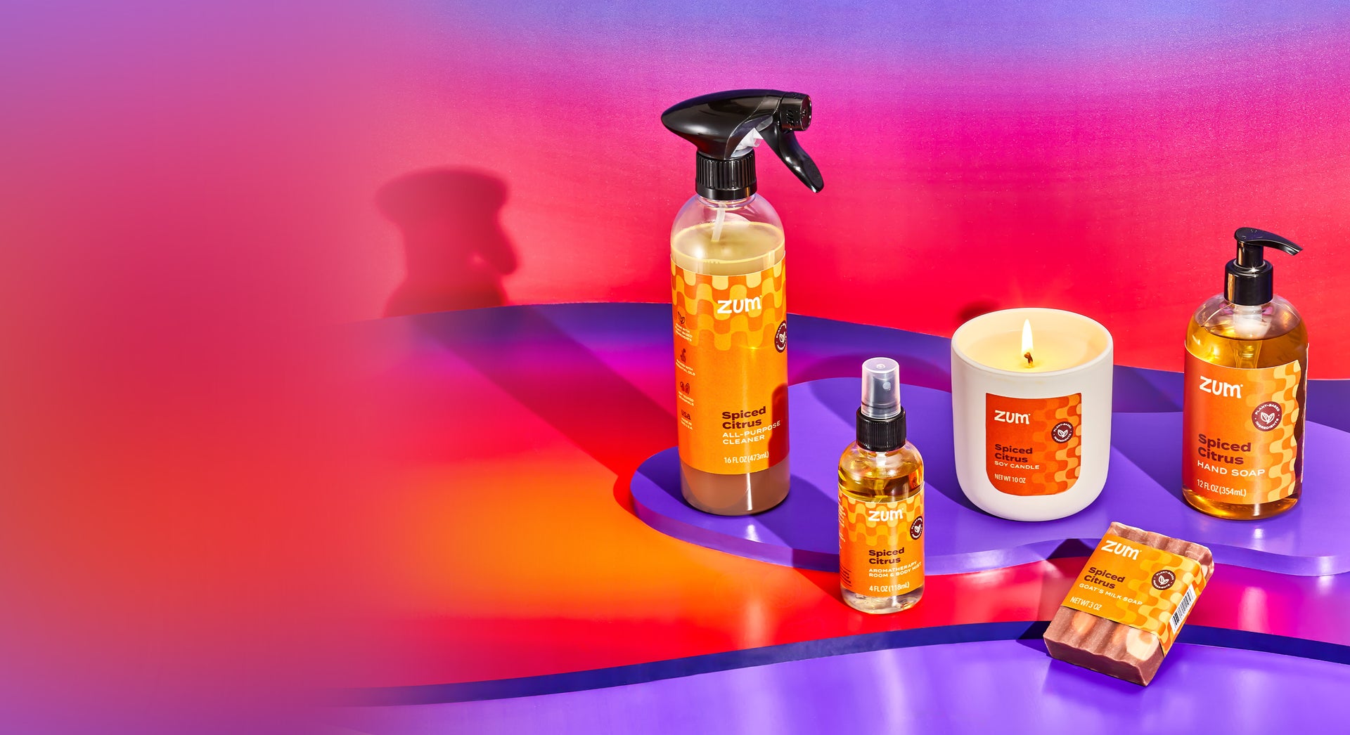 Spiced Citrus all-purpose cleaner bottle with a sprayer, room & body mist bottle with sprayer, soy candle, labeled bar soap, and hand soap bottle wit ha pump standing on purple and thermal gradient blob shaped risers.