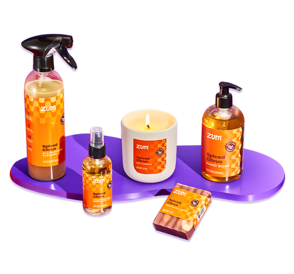 Spiced Citrus all-purpose cleaner bottle with a sprayer, room & body mist bottle with a sprayer, soy candle, bar soap, and hand soap bottle with a pump on a purple blob shaped riser.